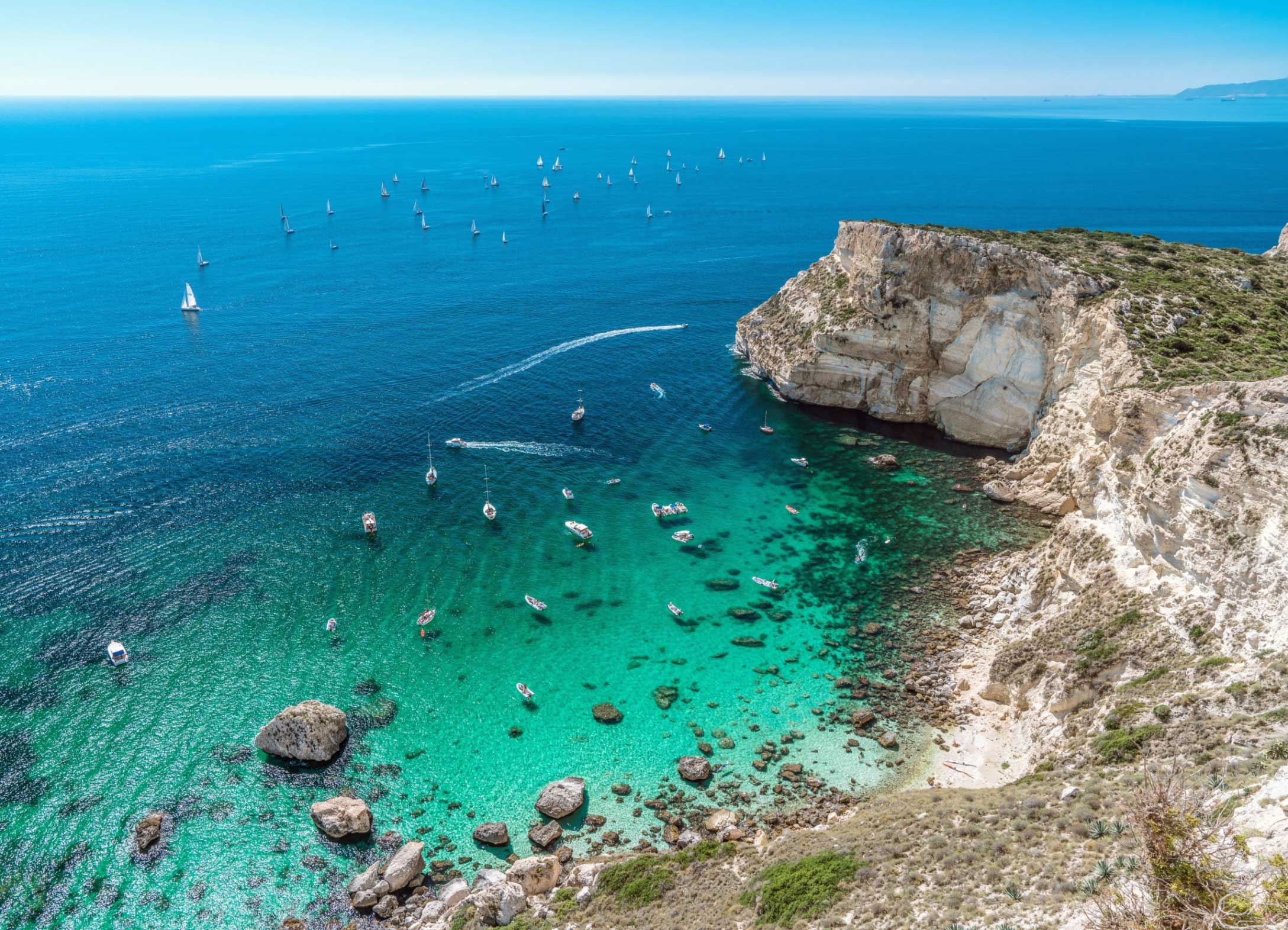Cala Fighera vue d'en haut : la falaise blanche à pic et les bateaux sur l'eau turquoise